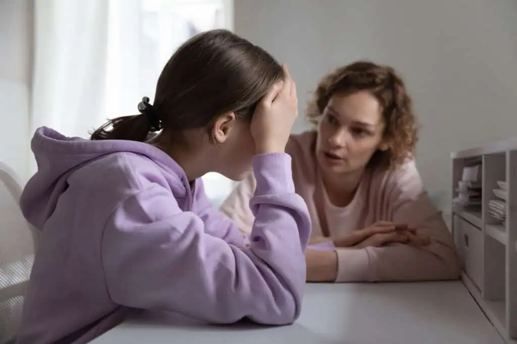 Two individuals sit at a white desk in a brightly lit room. The person on the left, wearing a purple hoodie, leans on the desk with their head resting in one hand—a classic sign your child doesnt respect you. The person on the right, in a pink sweatshirt, is talking to them.