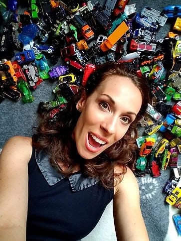 Tara Clark lies on the floor surrounded by numerous toy cars. She is smiling widely and appears to be taking a selfie. She has curly hair and is wearing a sleeveless black top with a collar. The toy cars are of various colors and types.