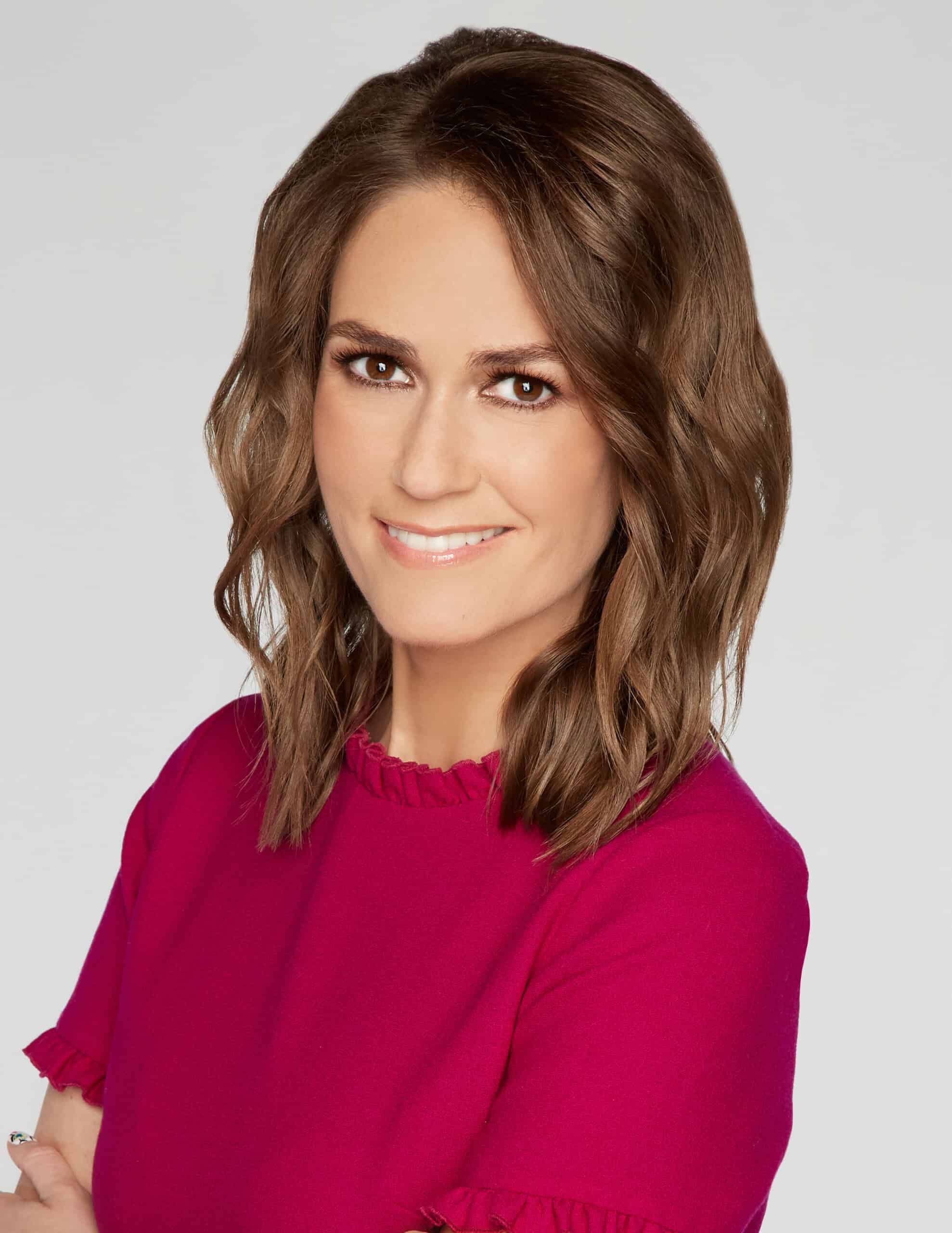 Jessica Tarlov with shoulder-length, wavy brown hair smiling while looking at the camera. They are wearing a bright pink top with ruffled edges. The background is plain white.