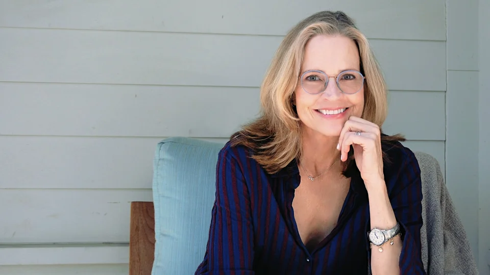 Janet Lansbury, wearing glasses and dressed in a dark blue shirt with red stripes, is sitting in a light-colored chair. She has her left hand resting on her chin and is smiling at the camera. She is wearing a watch on her left wrist.