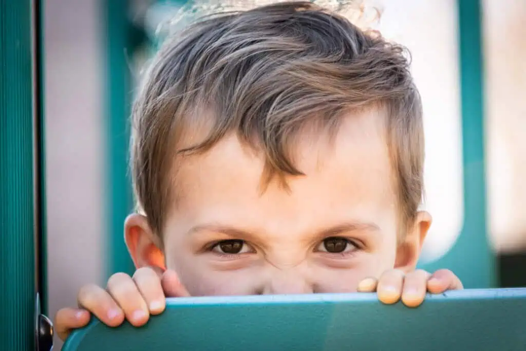 A bratty young child with light brown hair peeks over a teal-colored surface, squinting their eyes and making a playful face. The background is slightly blurred.