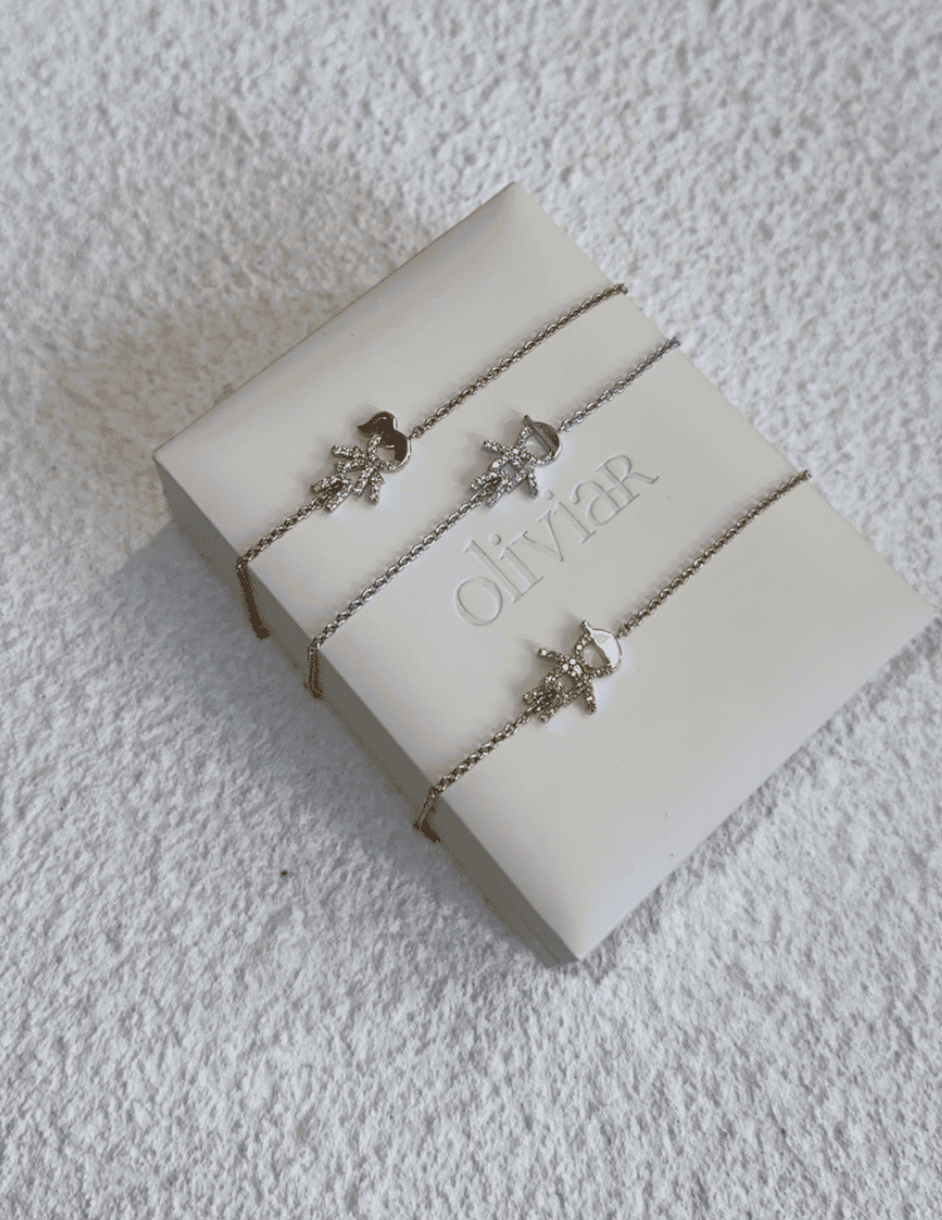 The image shows three delicate OliviaR bracelets, each featuring a small star charm, displayed on a white Olivia R branded box. The bracelets are in different metal tones: gold, silver, and rose gold. They are laid flat side by side on a textured white surface. Discover elegance with OliviaR.