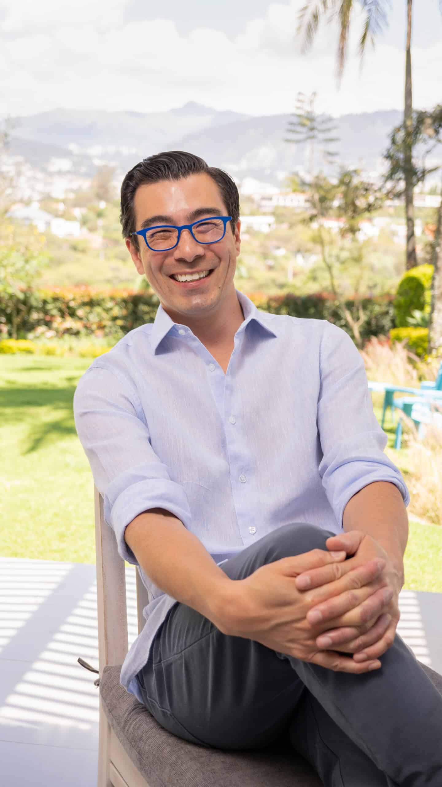 David Delisle wearing a light blue button-up shirt and dark pants sits outdoors on a chair. He is smiling, with his legs crossed and his hands resting on his knee. He is also wearing blue glasses. The background features a sunny landscape with greenery and hills.
