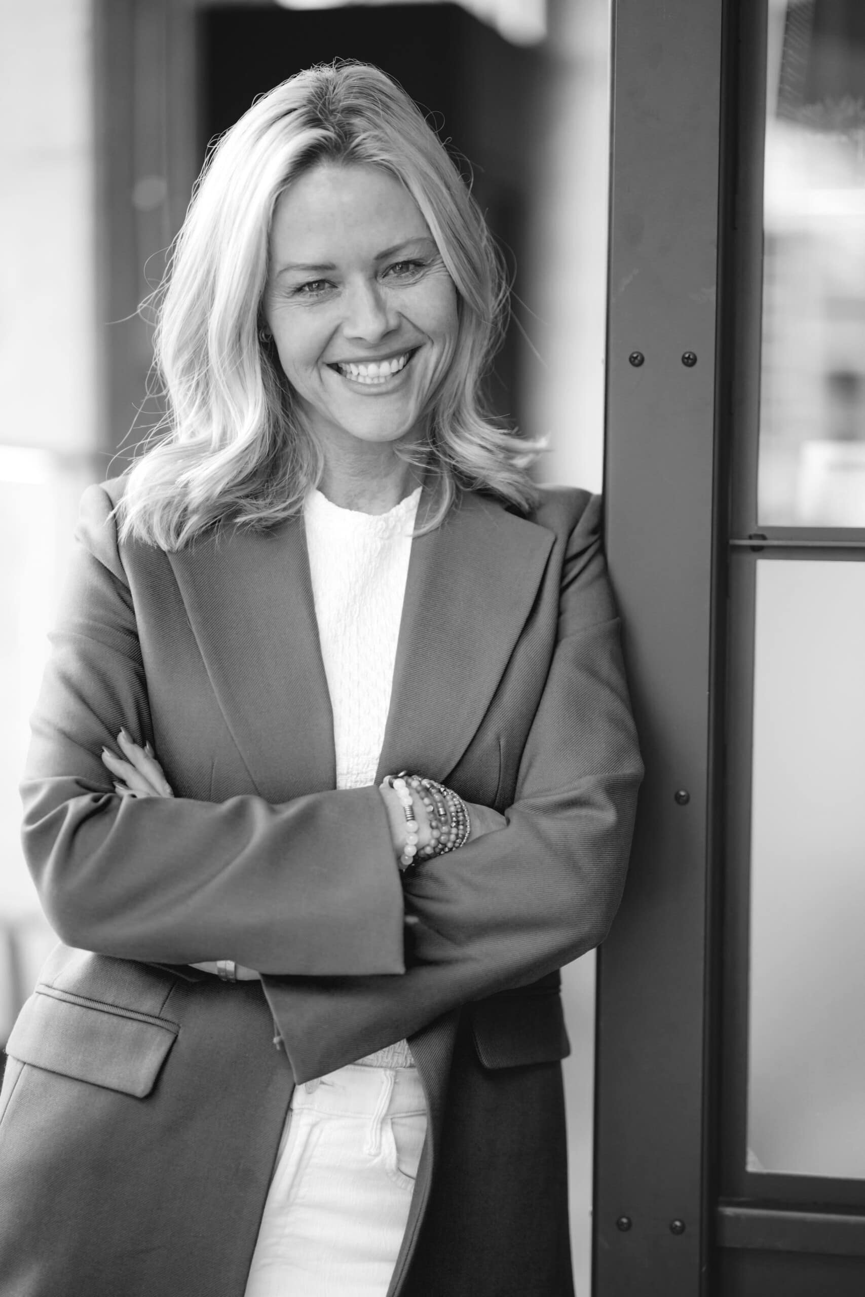 Black and white photo of a smiling woman, Kristin Andrus, with shoulder-length light hair, arms crossed, wearing a blazer over a white top and light-colored pants. She is standing against a door frame.