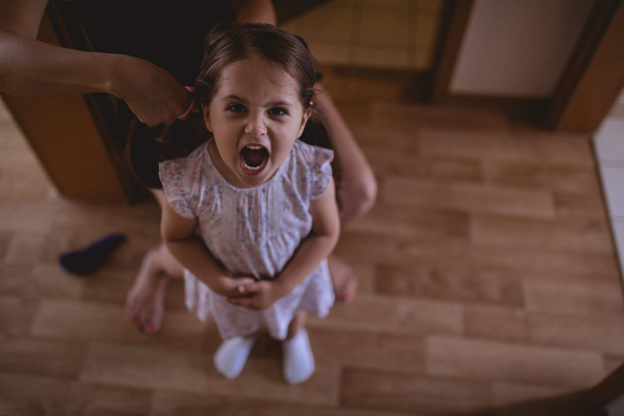 A young girl in a floral dress stands on a wooden floor with her mouth wide open, appearing to be shouting or yelling. An adult, partially visible, is behind her, styling the girls hair. One sock and a hairbrush are on the floor nearby—how to handle back talk evident in their dynamic.