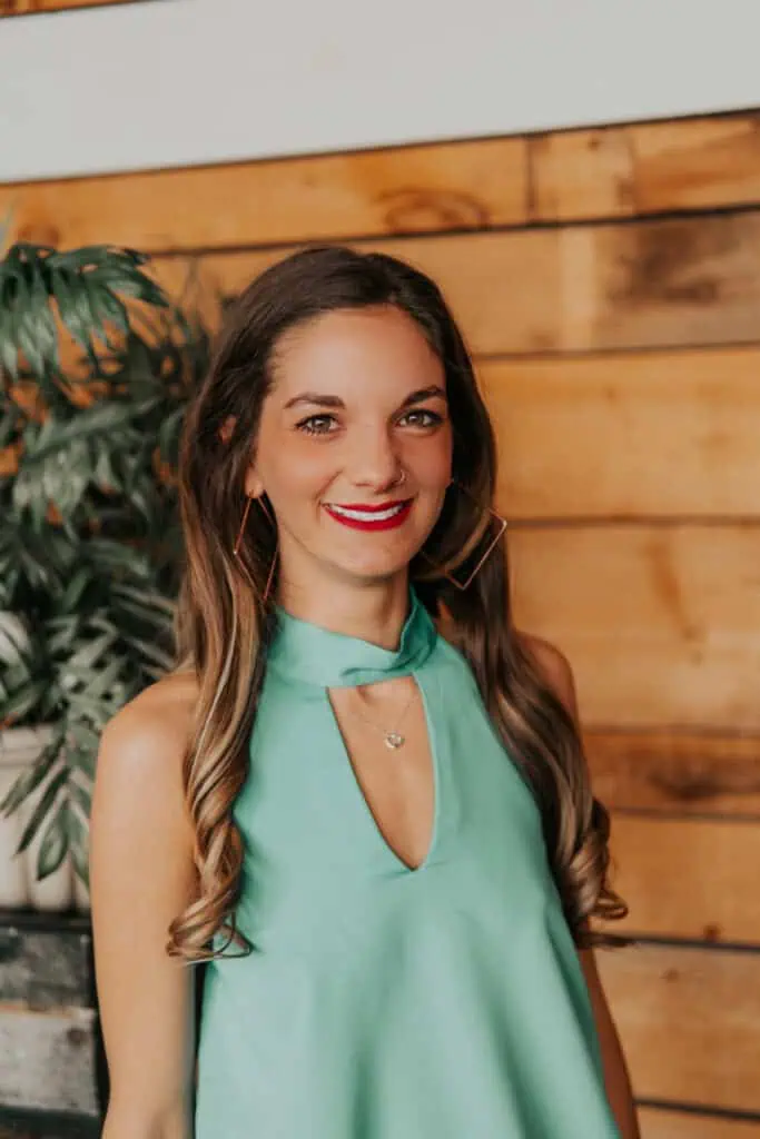 HeHe Stewart's headshot. She has long brown hair and is wearing a sleeveless green top. She is standing in front of a wooden wall. She is smiling and wearing large hoop earrings. There is a plant in the background.