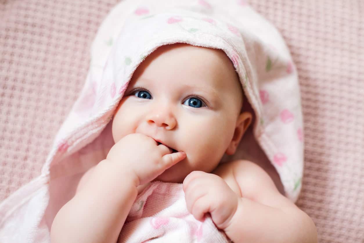 A baby girl wrapped in a soft, pink hooded towel adorned with heart patterns gazes forward, one hand in their mouth. Lying on a textured pink surface.