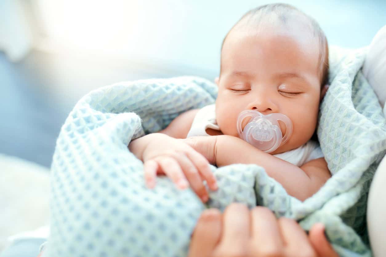 A sleeping baby boy with a pacifier rests in a light blue blanket, nestled securely in someones arms. The softly lit background highlights this serene scene, inviting thoughts of tender moments.