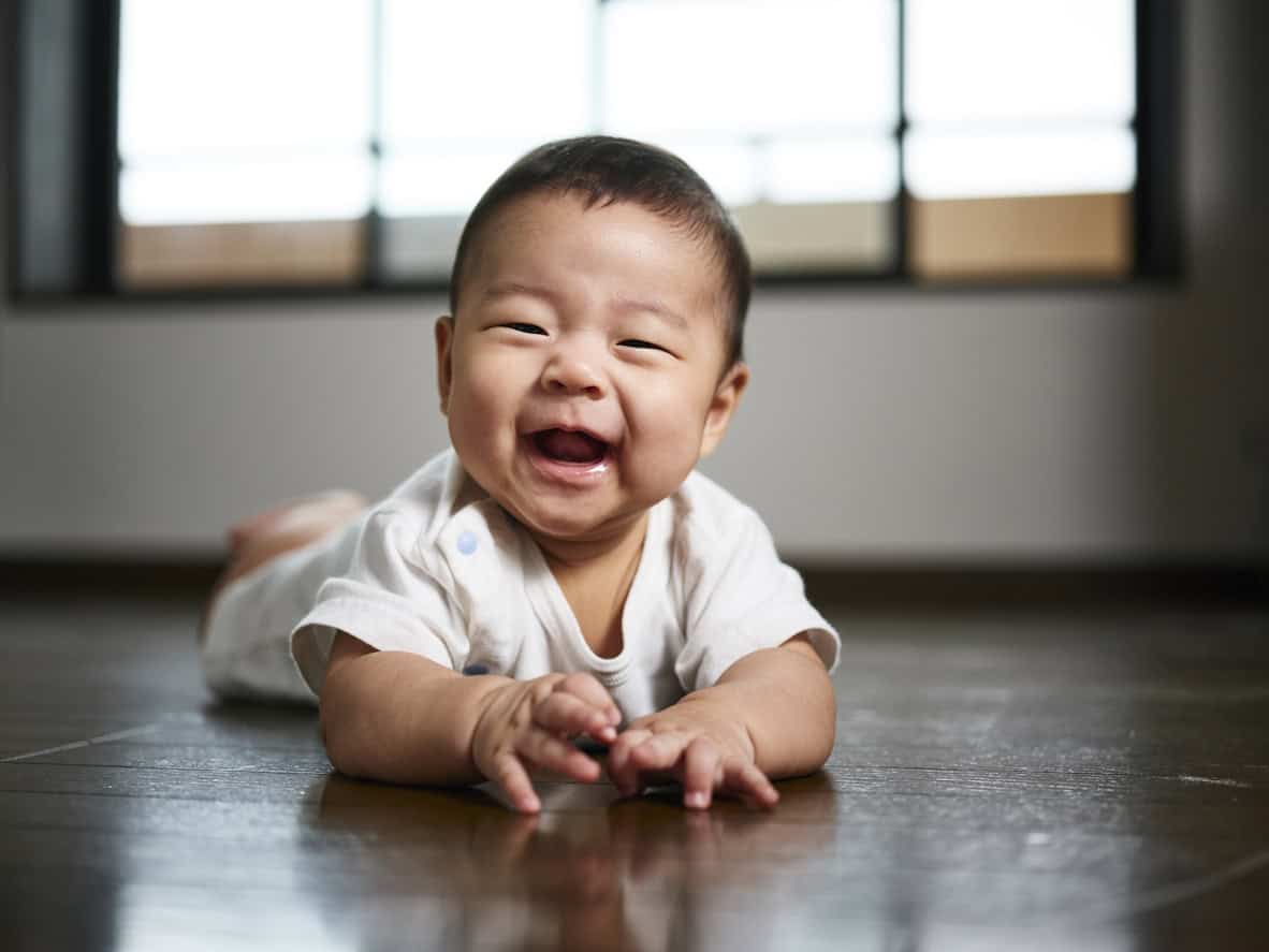 A boy baby wearing a white onesie is lying on his stomach on a wooden floor, smiling or laughing. In the background, large windows with blurred details add warmth to the scene.