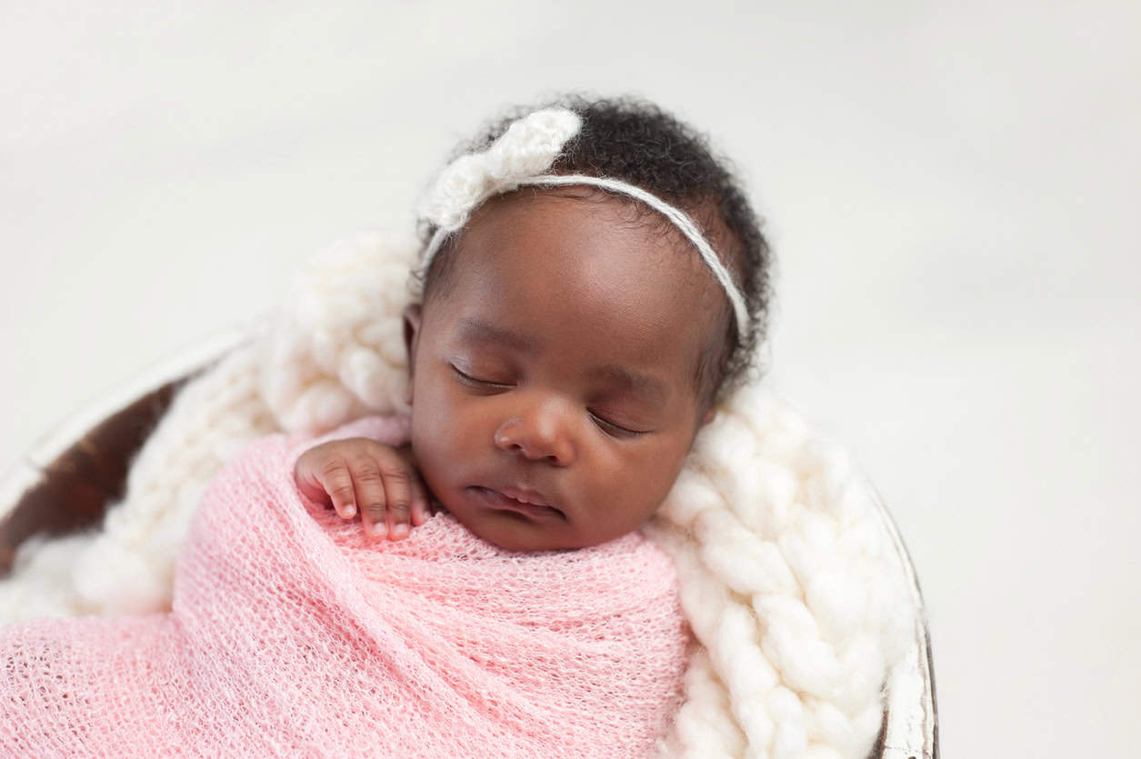 A sleeping baby girl wrapped in a pink blanket lies on a soft white knitted fabric. She wears a white headband with a small bow. The background is softly blurred, creating a peaceful atmosphere.