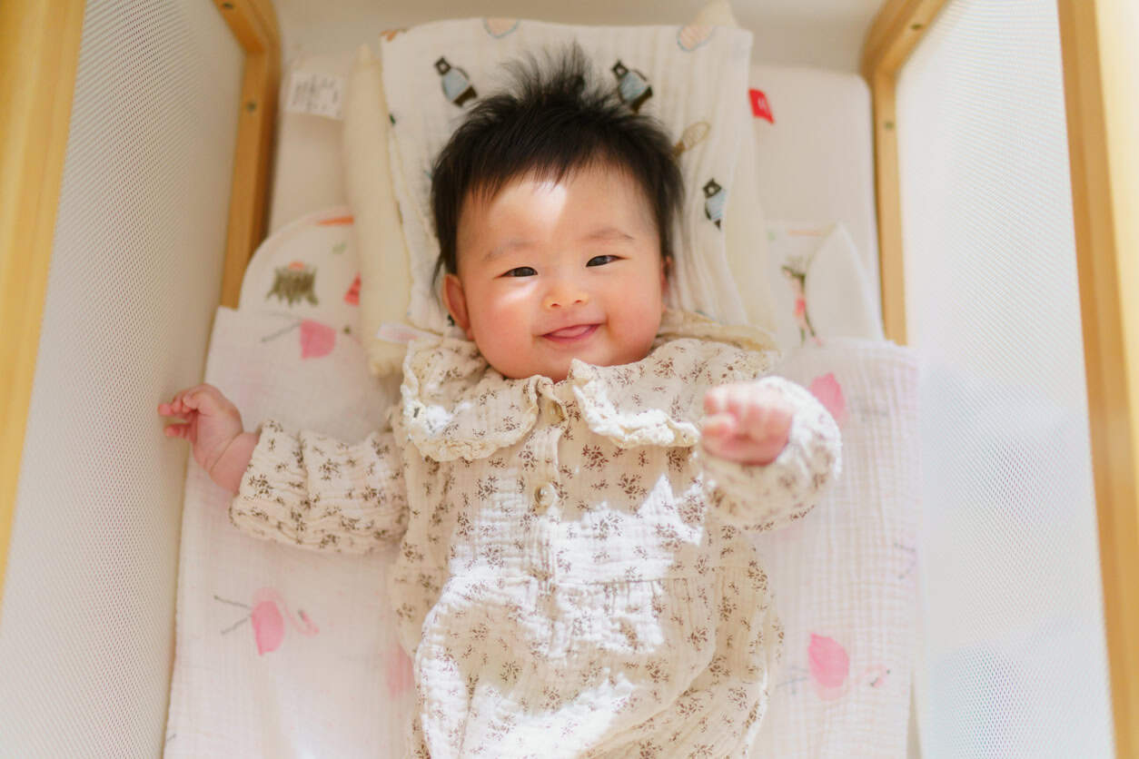 A baby girl with dark hair smiles while lying on her back in a crib. The baby girl is wearing a patterned cream-colored outfit, and the crib is lined with soft, patterned bedding.