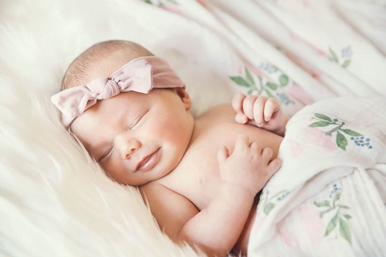 A sleeping newborn baby girl is covered with a floral-patterned blanket. She wears a pink bow headband and rests peacefully on a fluffy white surface.