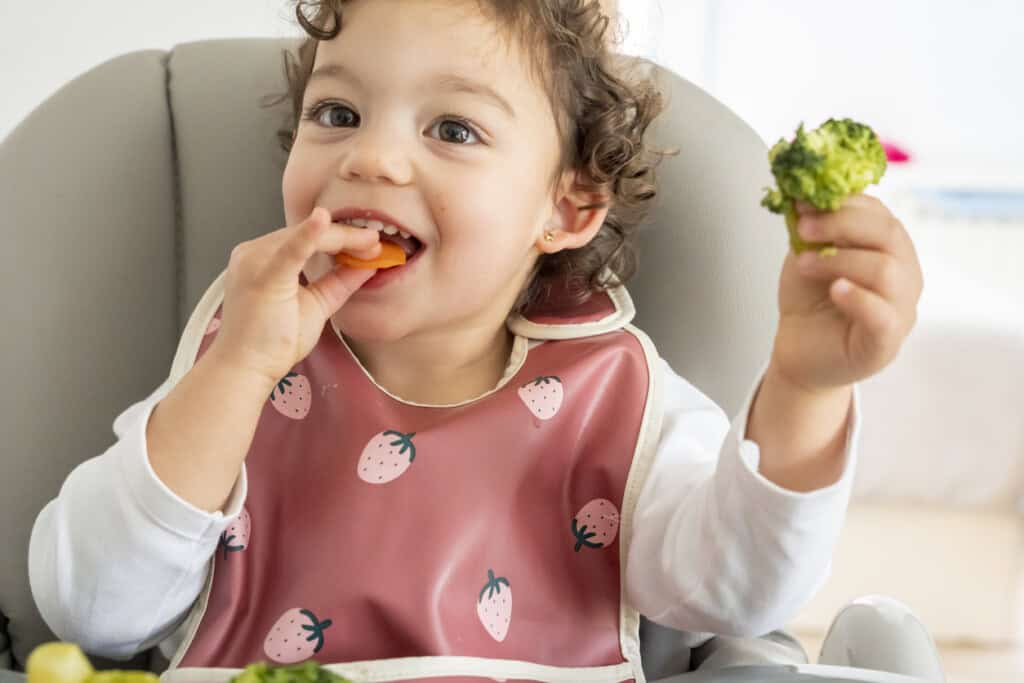 A young girl with curly hair sits in a high chair, wearing a bib with strawberry patterns. The child is holding a piece of broccoli in one hand and a carrot in the other, appearing to eat the carrot. A plate with more food is in front of them.
