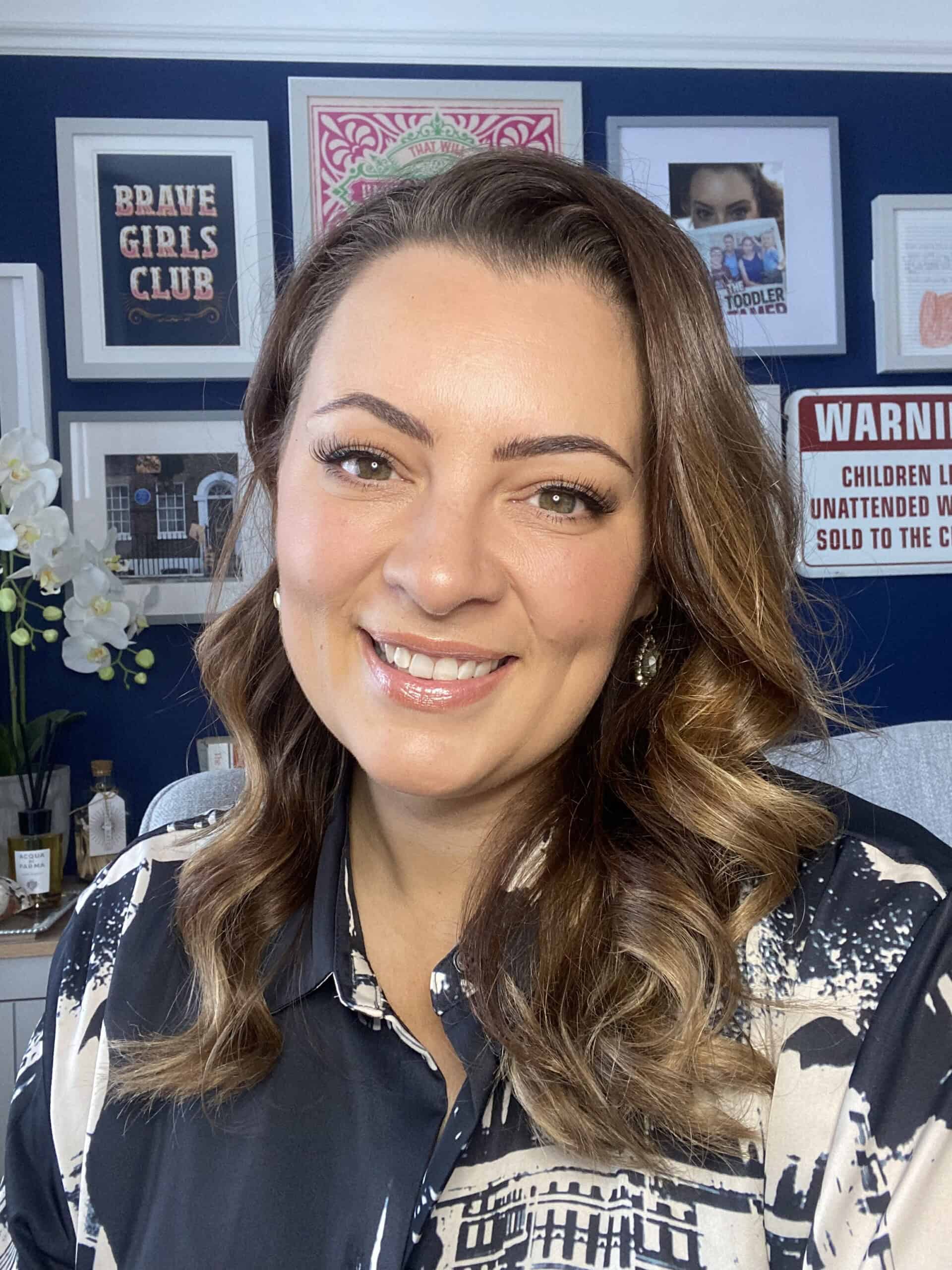 A woman with wavy brown hair smiles at the camera in a room with a navy blue wall decorated with framed pictures and signs. She wears a patterned shirt and pearl earrings. White flowers are visible in the background.