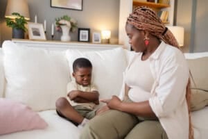 Angry Black Mother sitting down with Her Child Son at Home while he is pouting on the couch.
