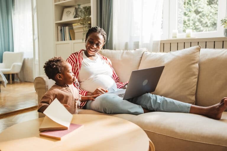 Pregnant young woman working on laptop while spending time with her little son at home.