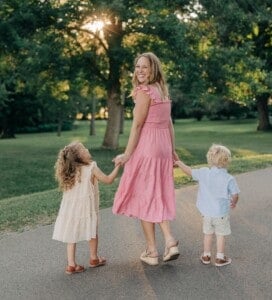 Young woman in pink dress holding hands with two kids on a park path.